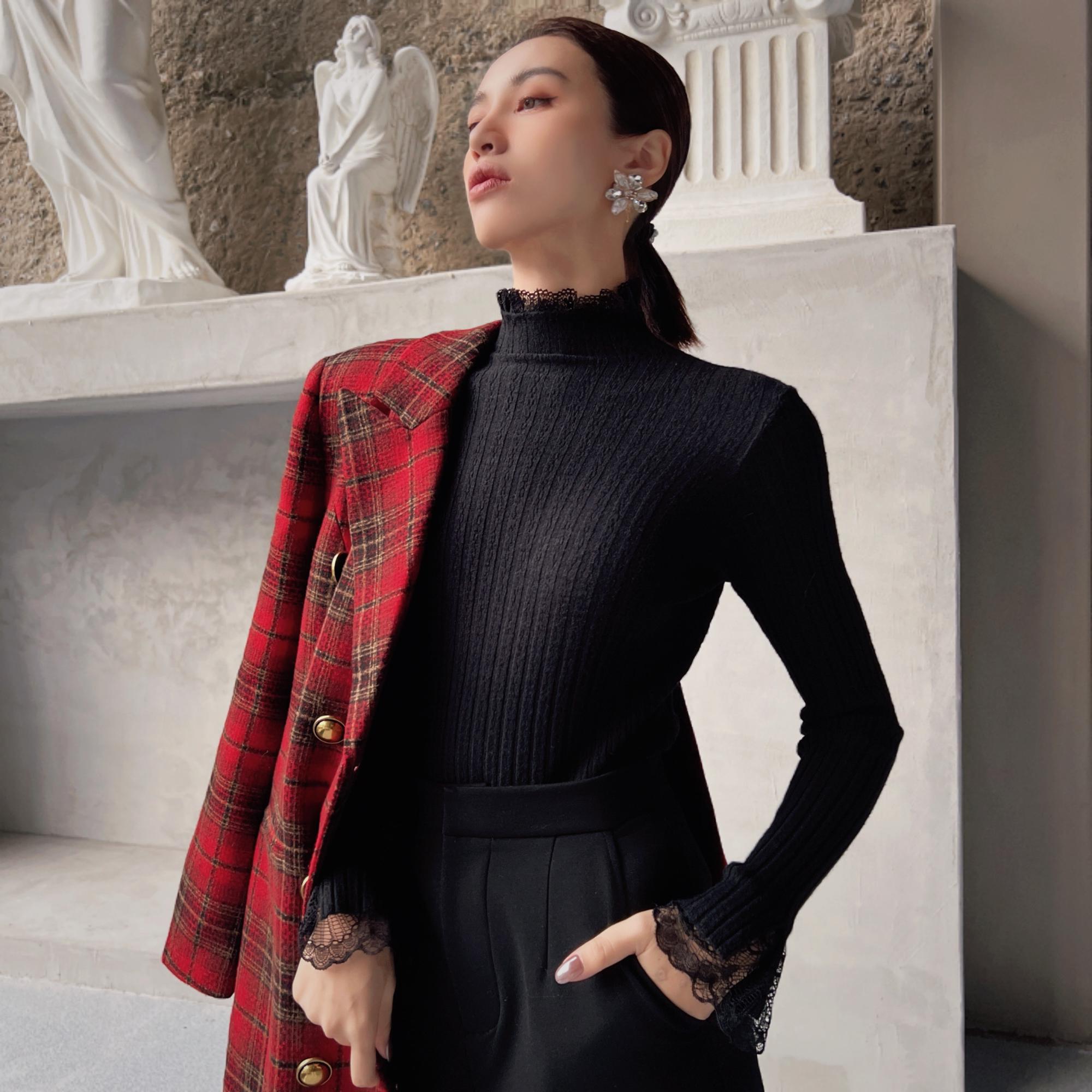 A woman wearing a black sweater and a red plaid jacket stands in front of a stone wall with statues.