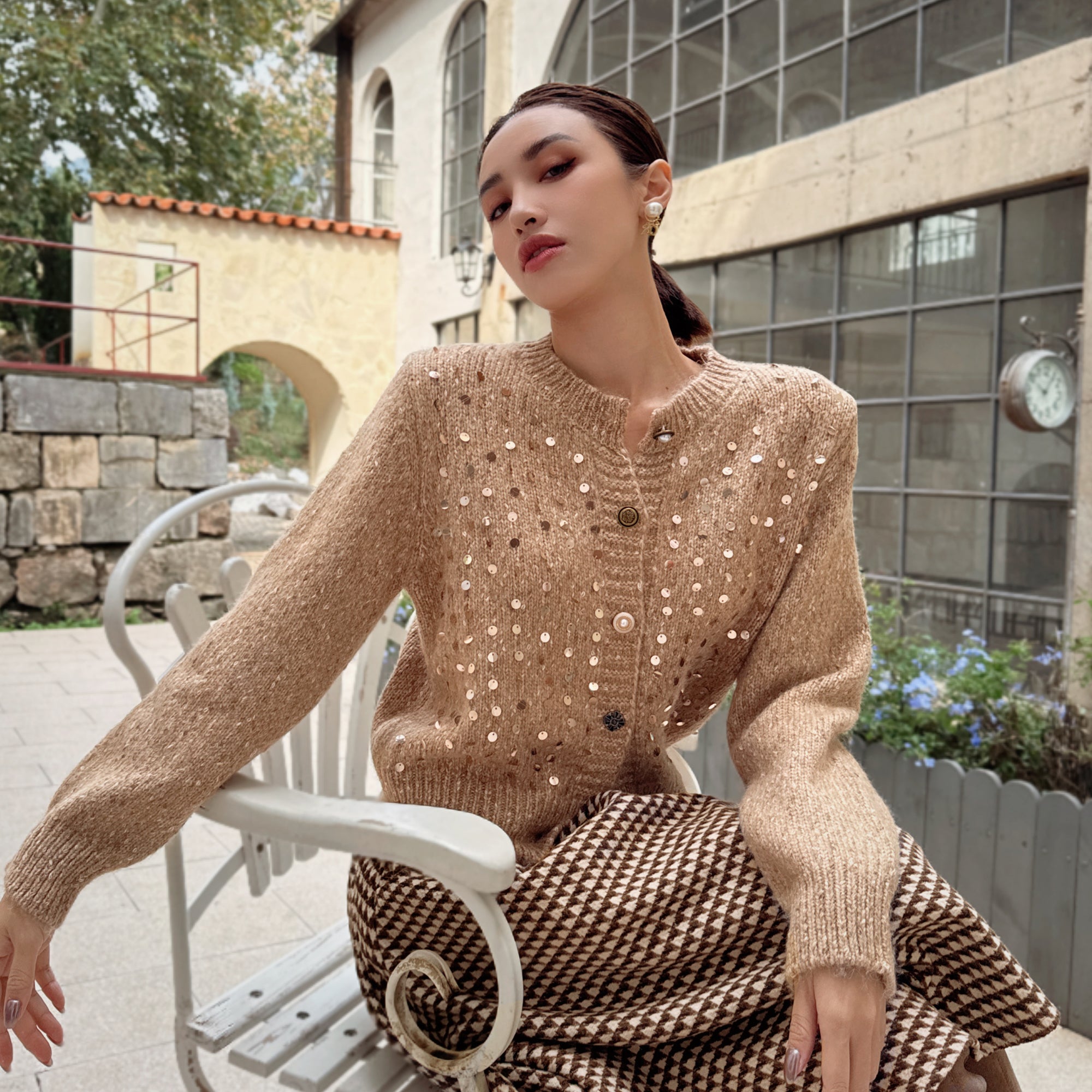 A woman wearing a beige sweater with white polka dots and a black and white checkered skirt is sitting on a white bench in front of a building.