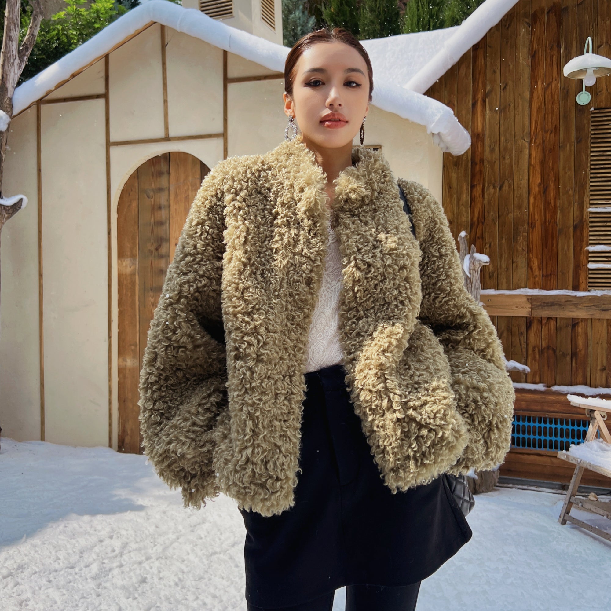 A woman wearing a beige fur coat stands in front of a wooden cabin with snow on the roof.