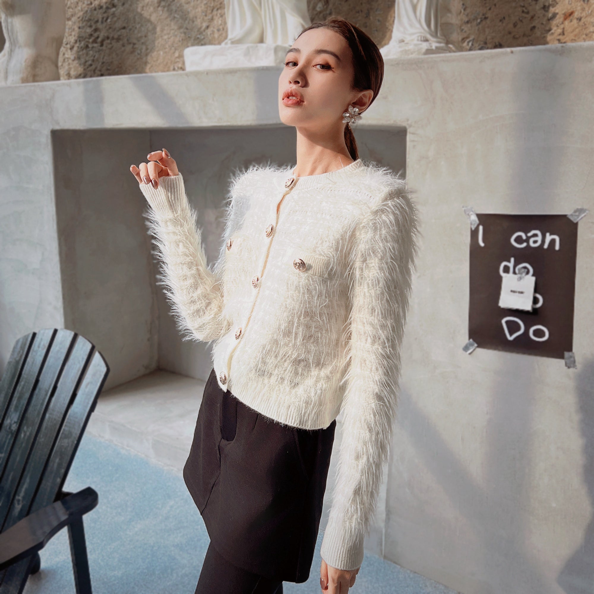 A woman wearing a white furry cardigan and black skirt stands in front of a concrete wall with a sign that says "I can do anything".