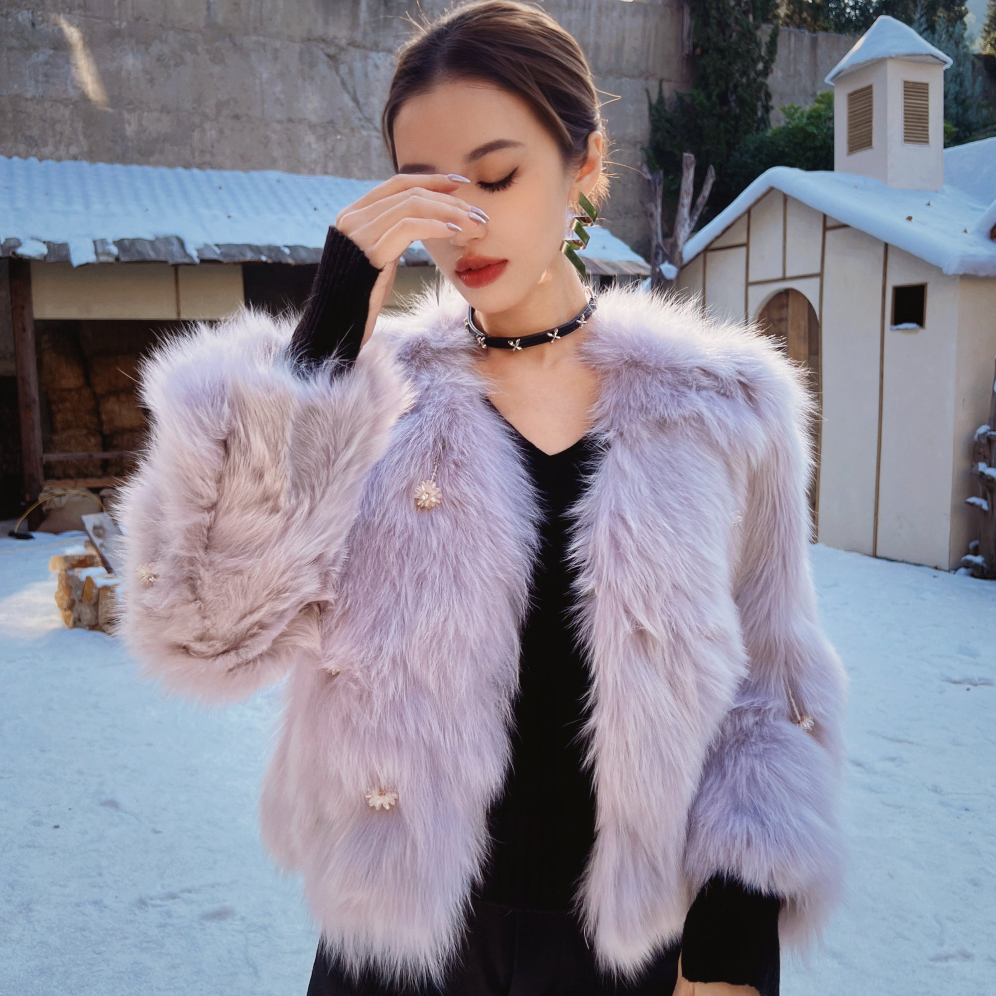 A woman wearing a pink fur coat stands in front of a snowy building, with her hand covering her face.
