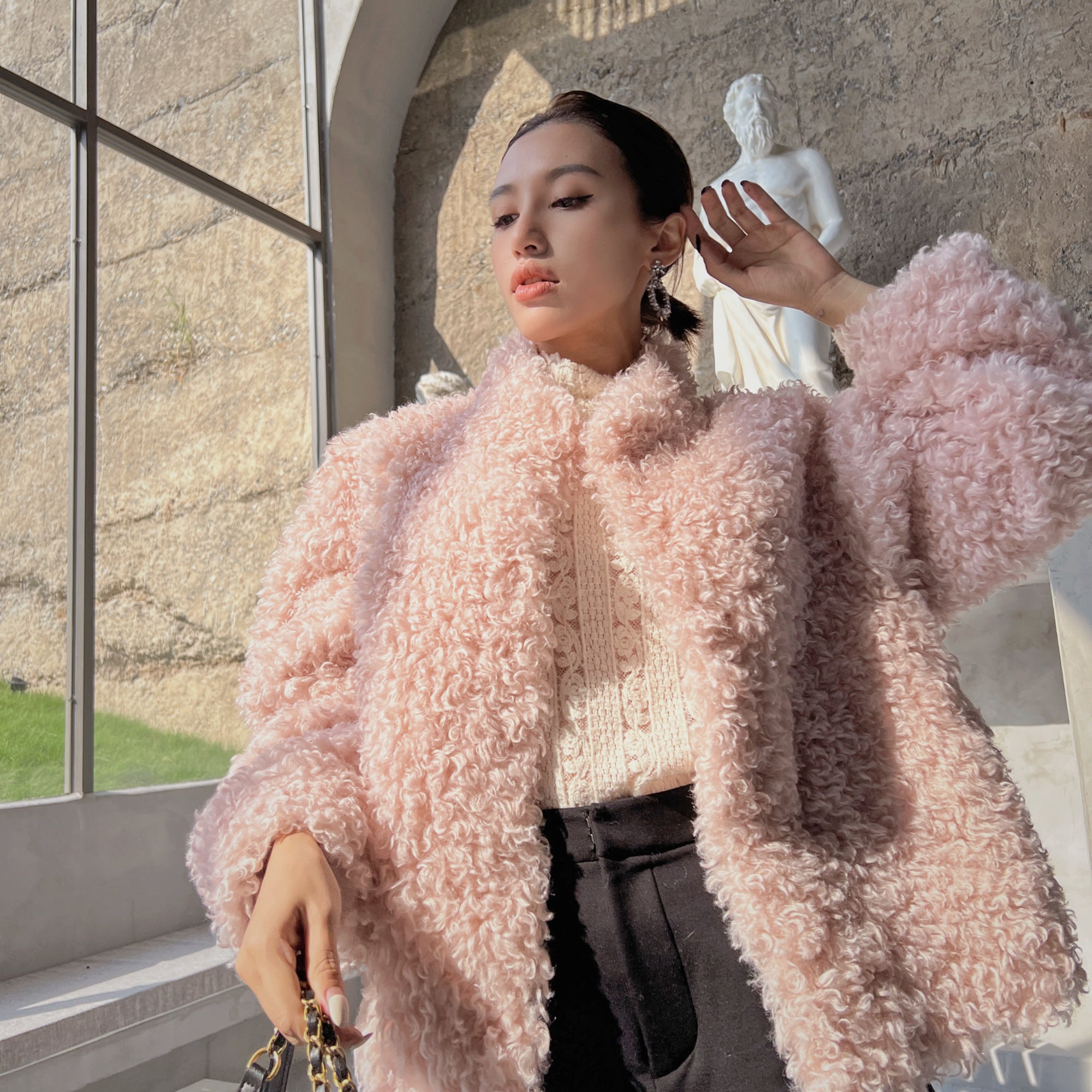 A woman wearing a pink fluffy coat stands in front of a window, with a statue visible in the background.