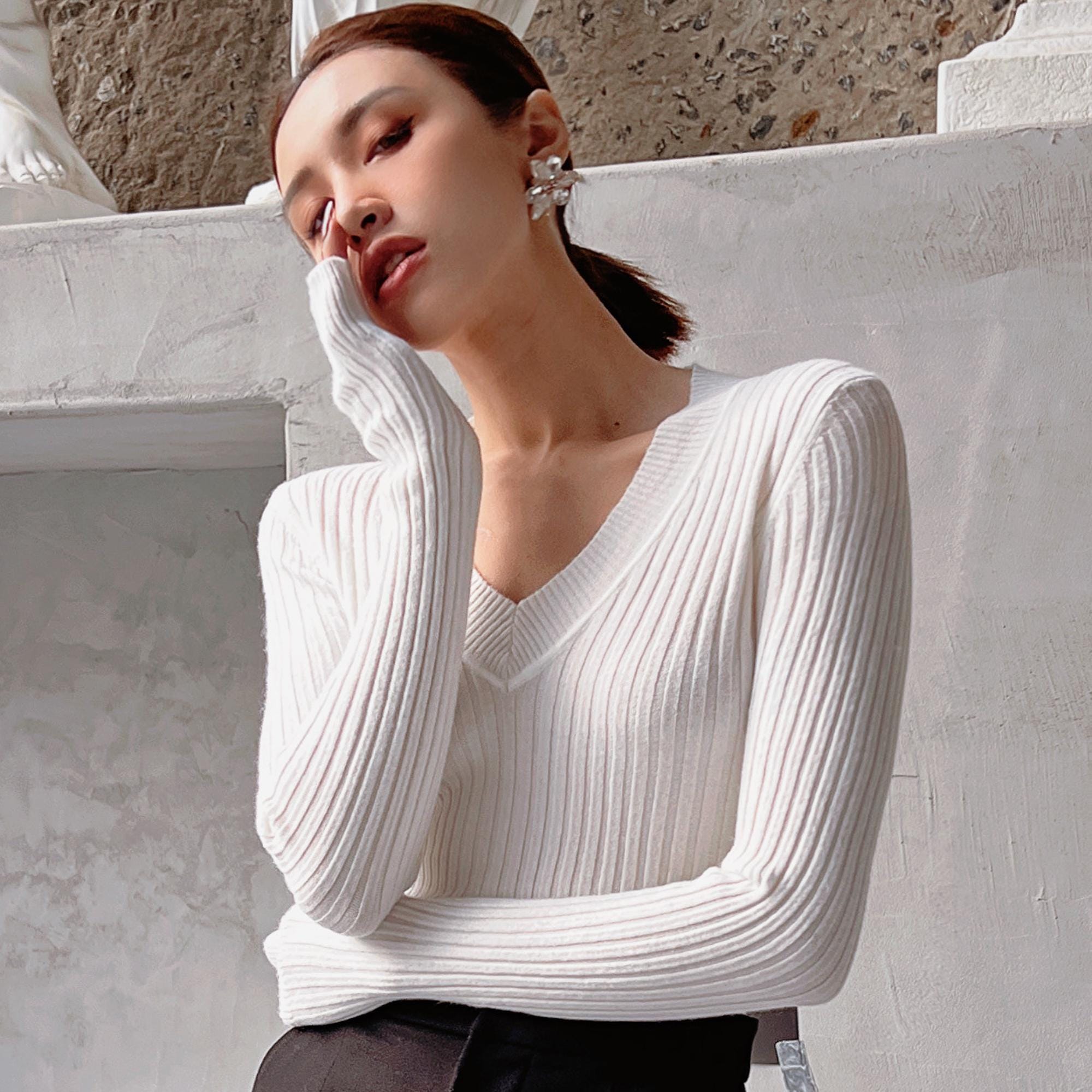 A woman wearing a white sweater and earrings is posing for a photograph, with her hand on her face.