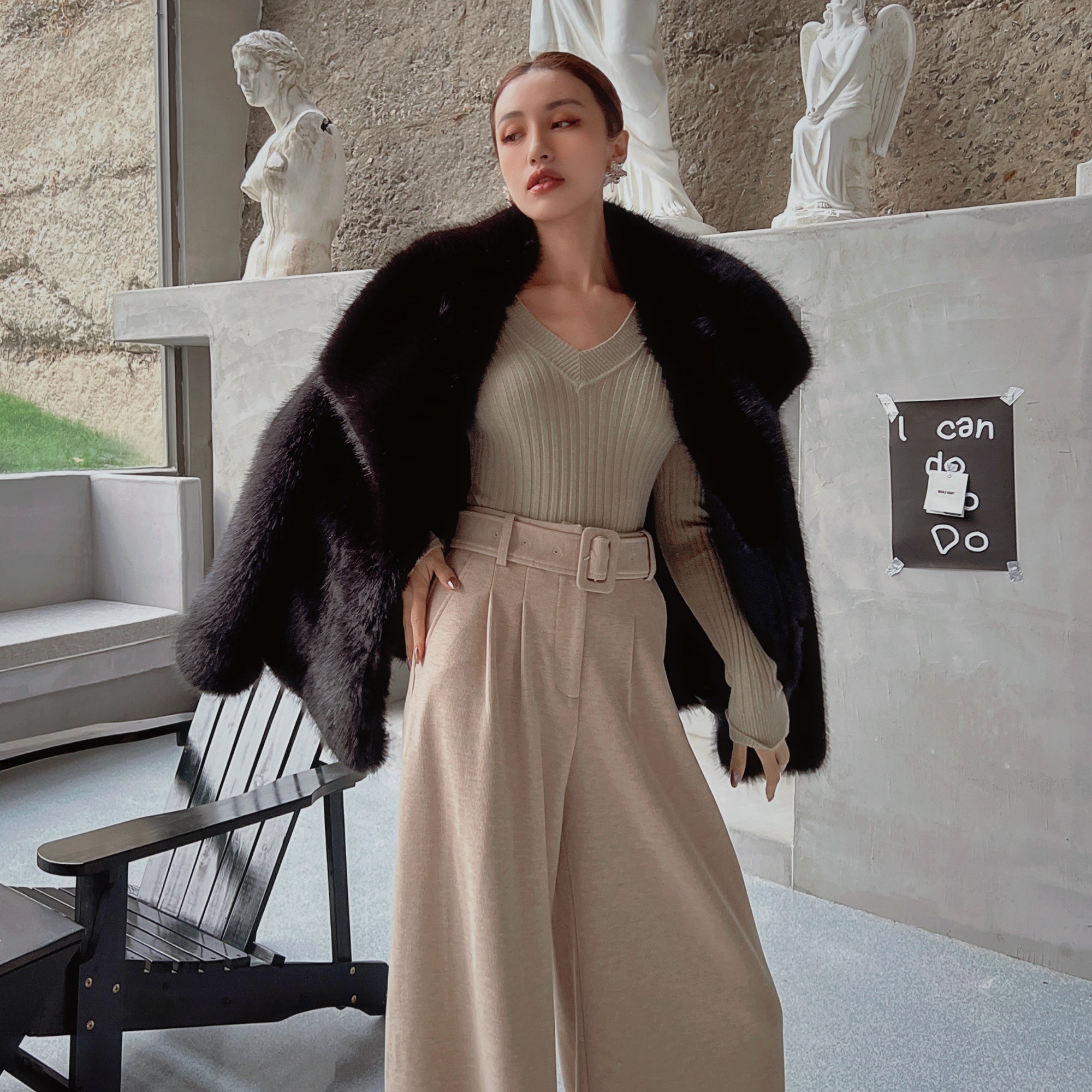 A woman wearing a black fur coat, beige sweater, and wide-leg pants stands in an indoor setting with a stone wall and statues in the background.