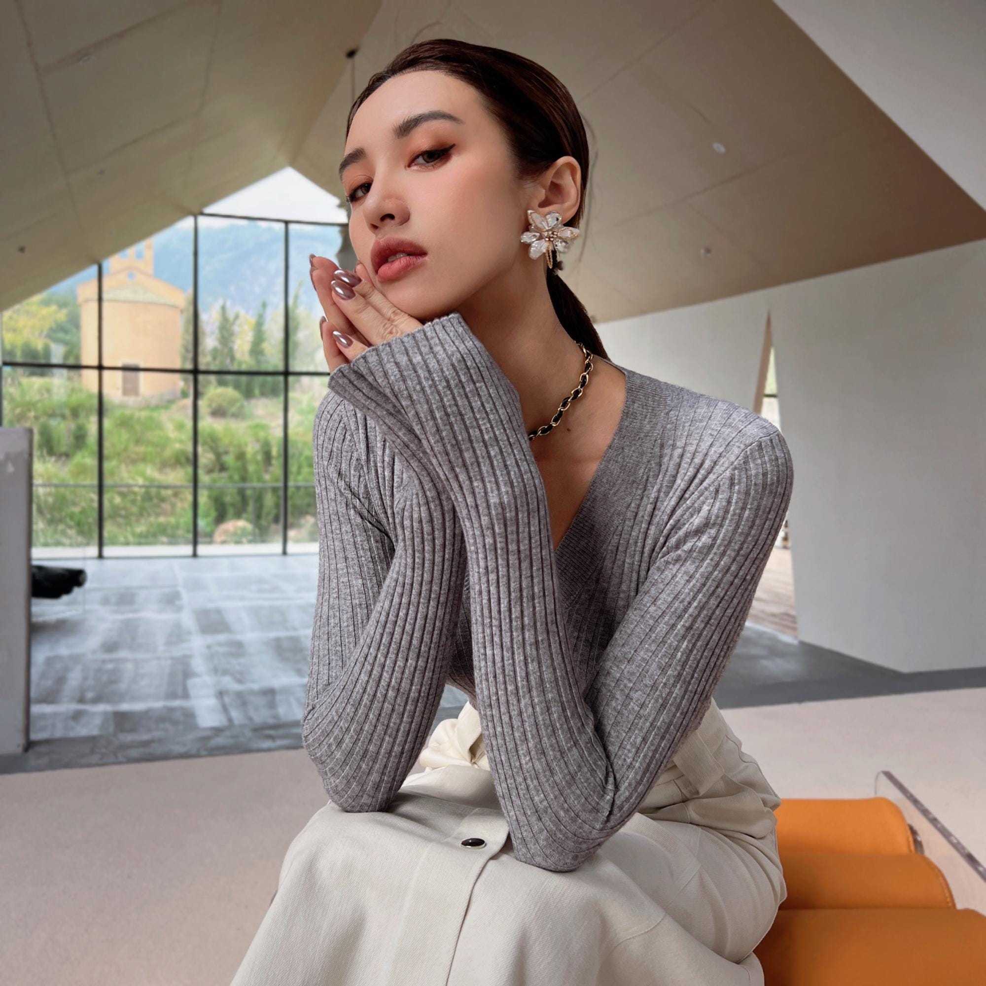 A woman wearing a gray sweater and white pants sits on a bench, looking pensive with her hands on her face.