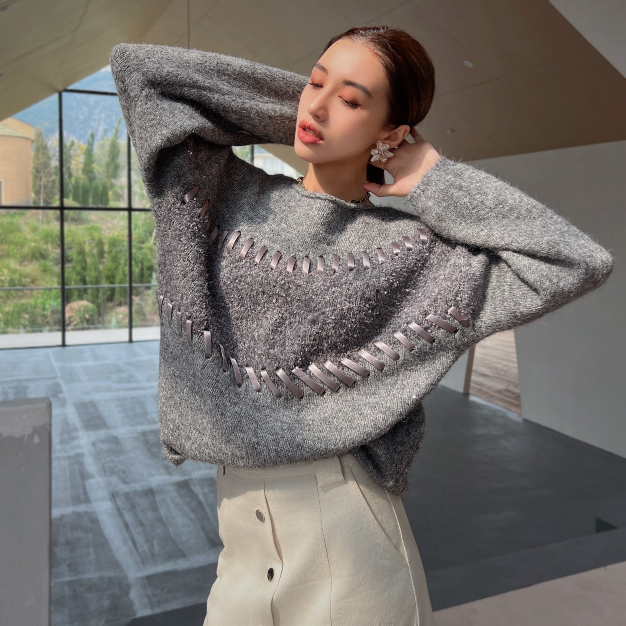 A woman wearing a gray sweater with a unique pattern stands in a room with large windows overlooking a natural landscape.