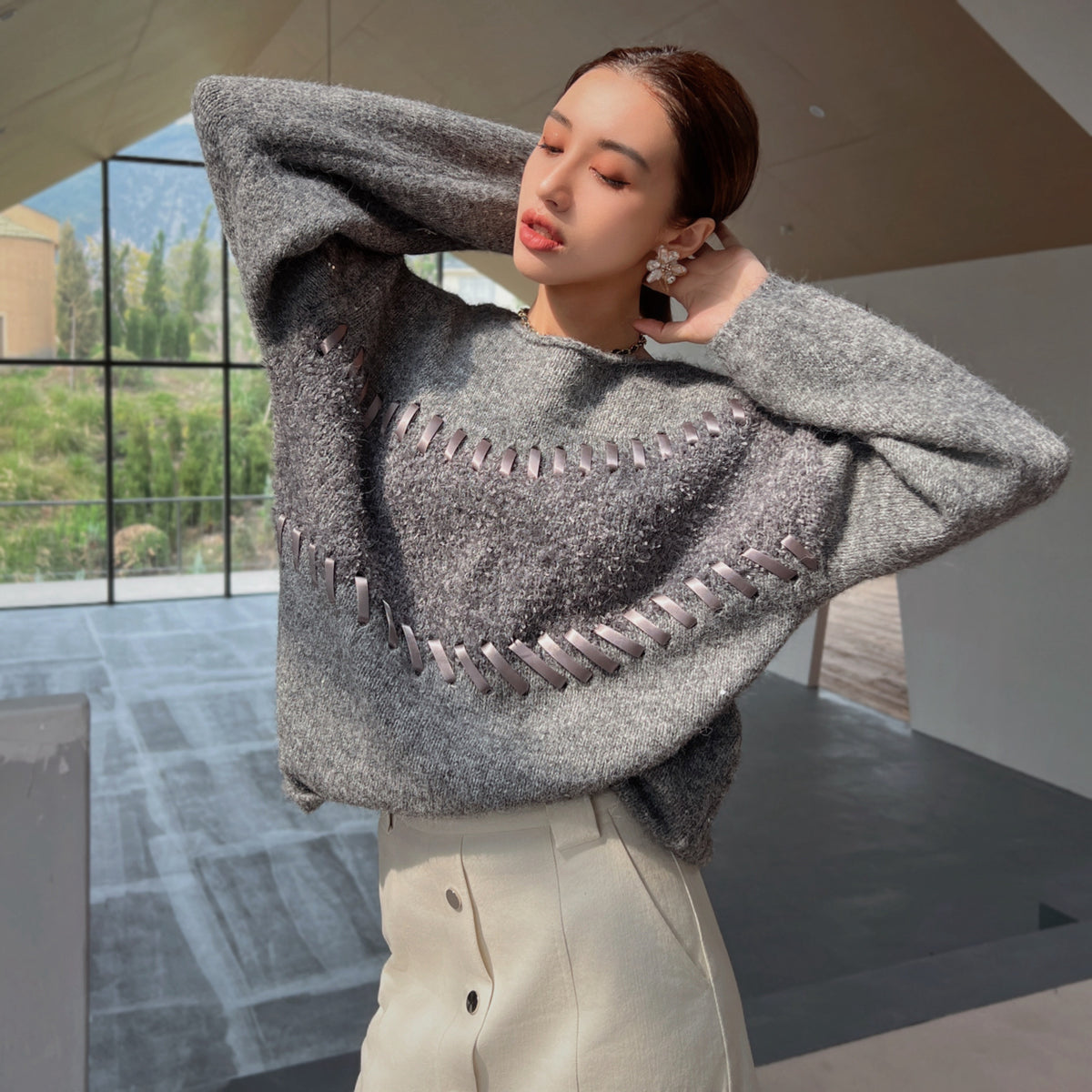A woman wearing a gray sweater with a unique pattern stands in a room with large windows overlooking a natural landscape.