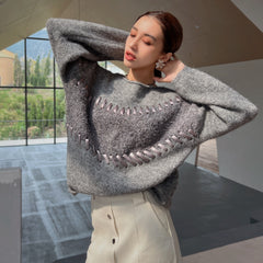 A woman wearing a gray sweater with a unique pattern stands in a room with large windows overlooking a natural landscape.