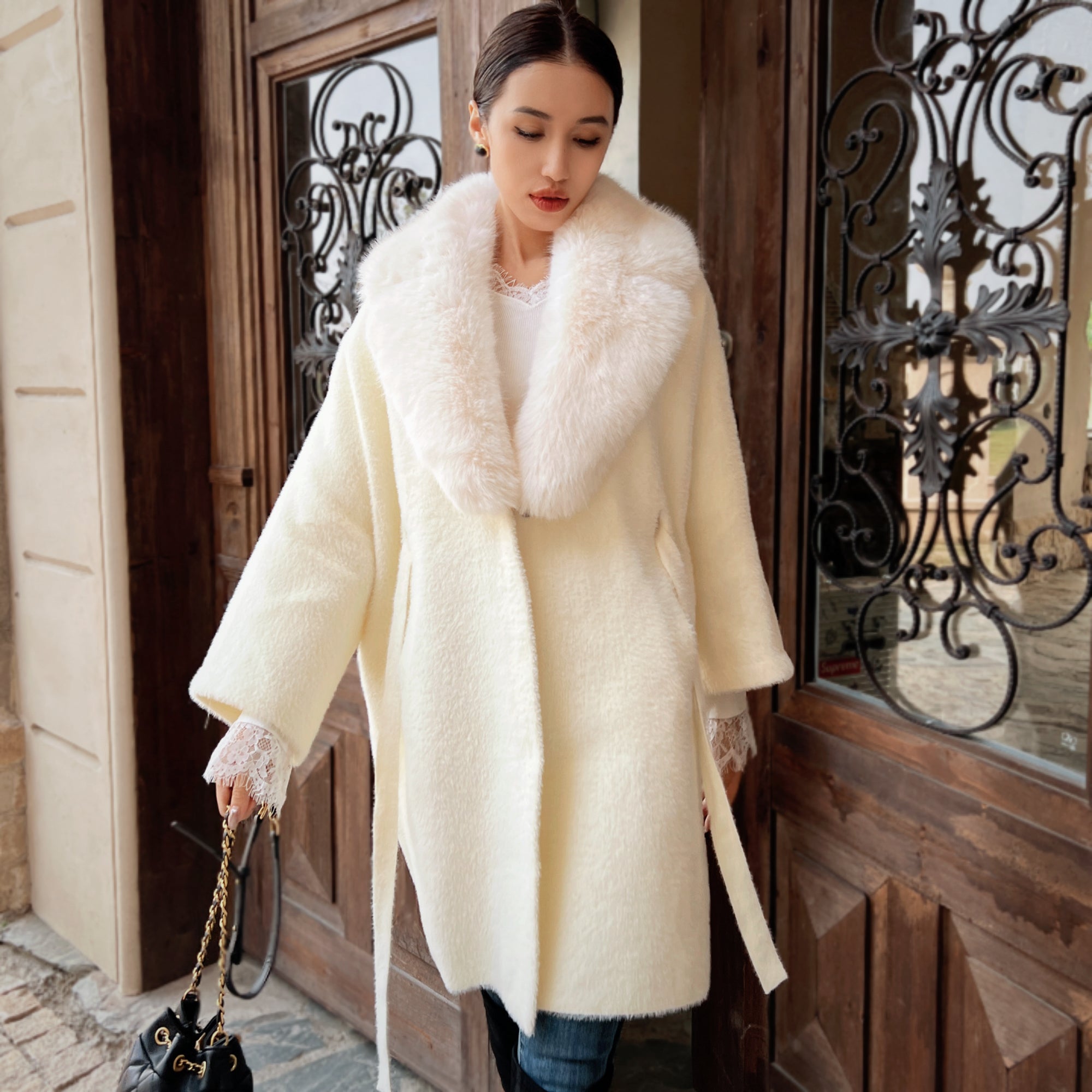 A woman wearing a white fur coat and carrying a black purse is standing in front of a wooden door.