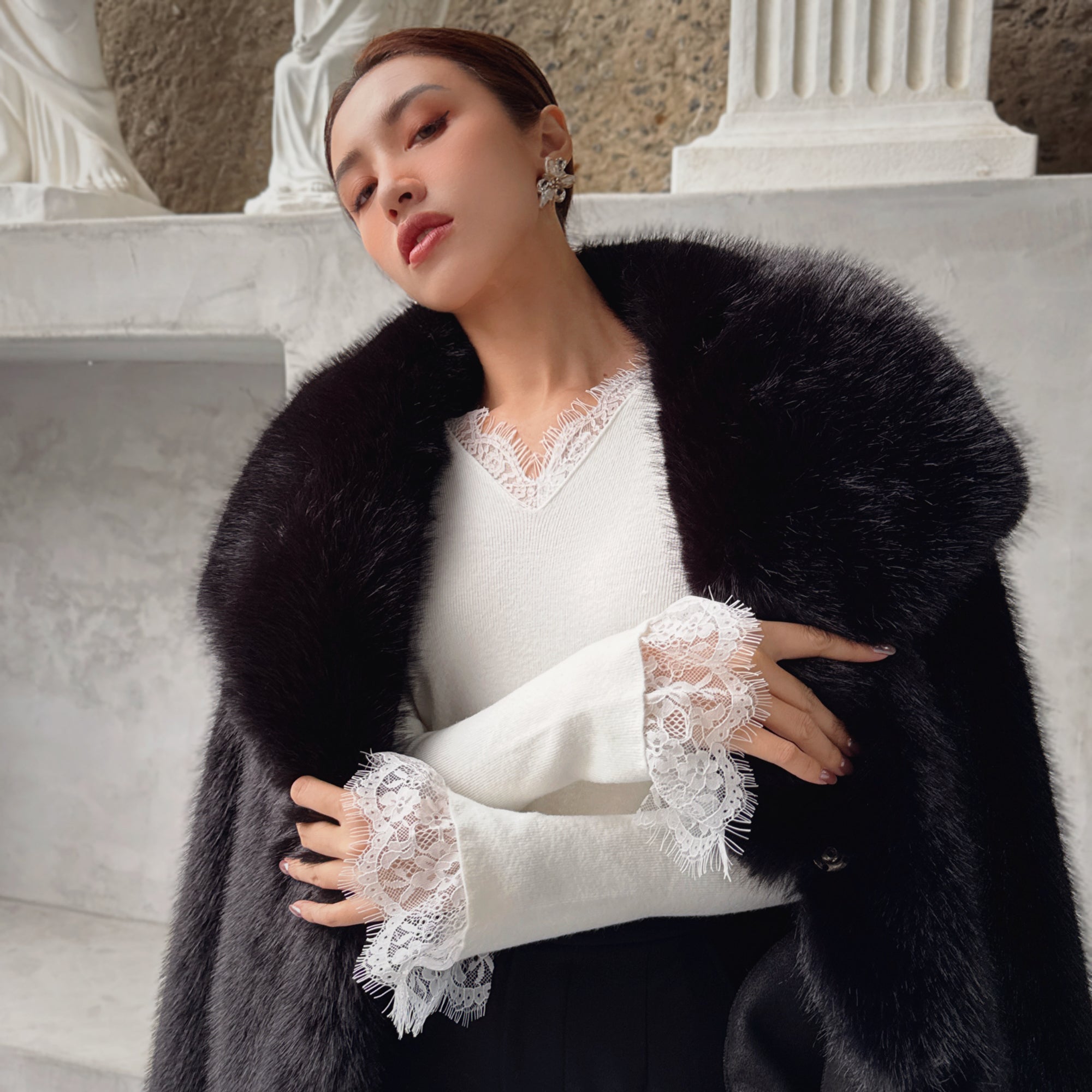 A woman wearing a white blouse with lace sleeves and a black fur coat stands in front of a stone wall.