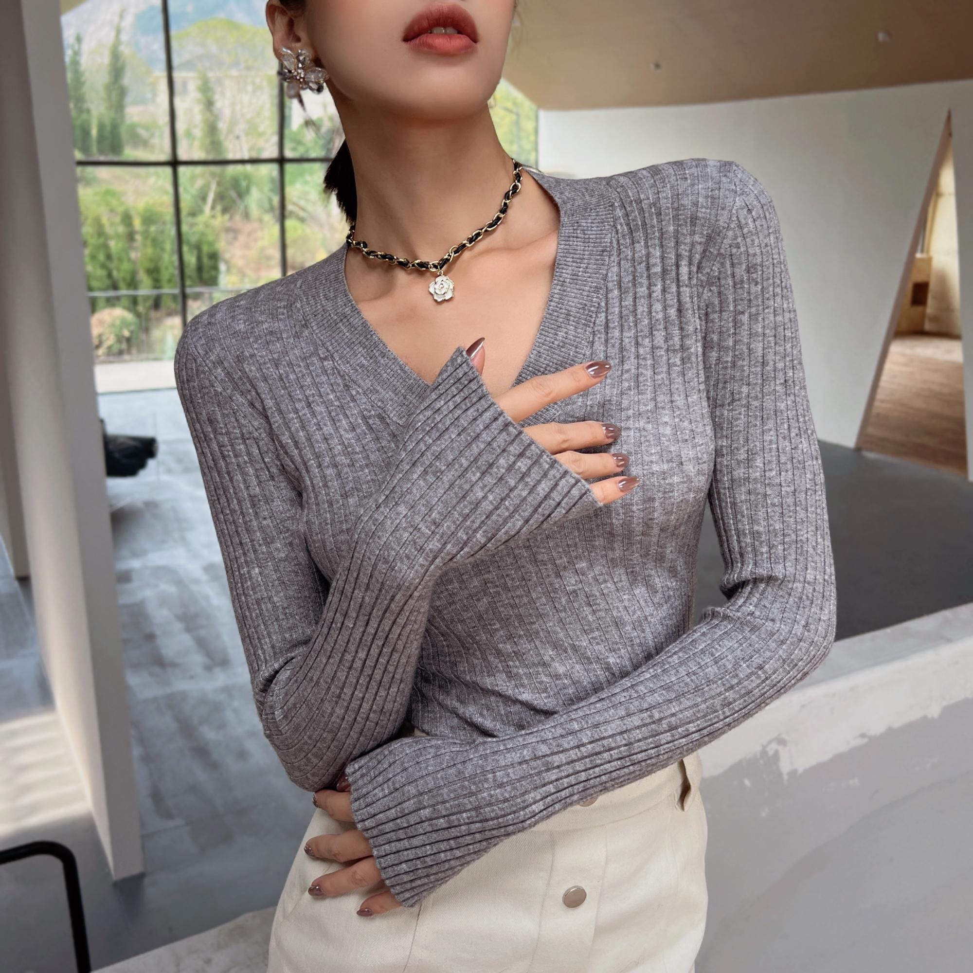 A woman wearing a gray sweater and white pants stands in a room with a window, her hands tucked into her pockets.