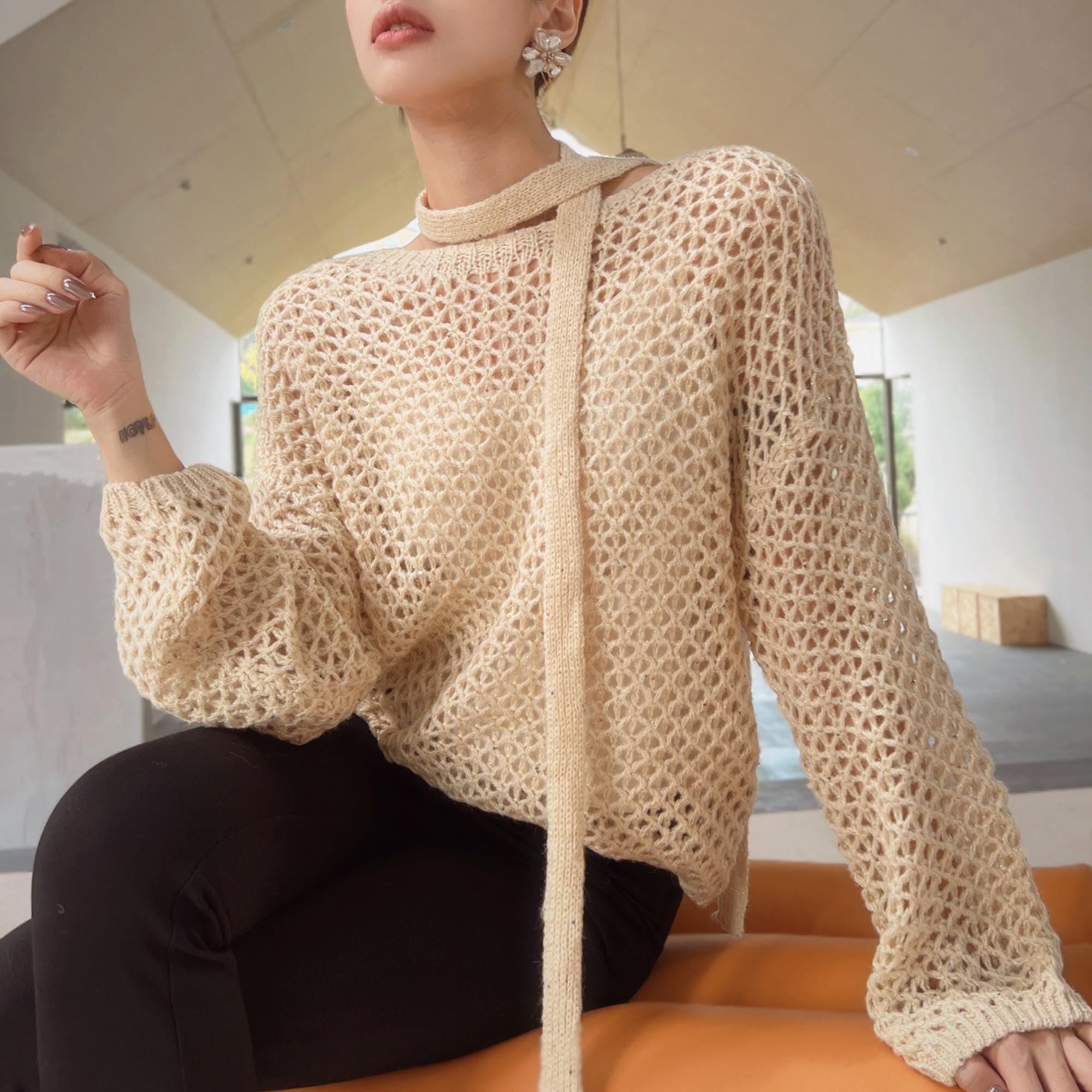 A woman wearing a beige crocheted sweater with a high collar and long sleeves, sitting on a bench in an indoor setting.