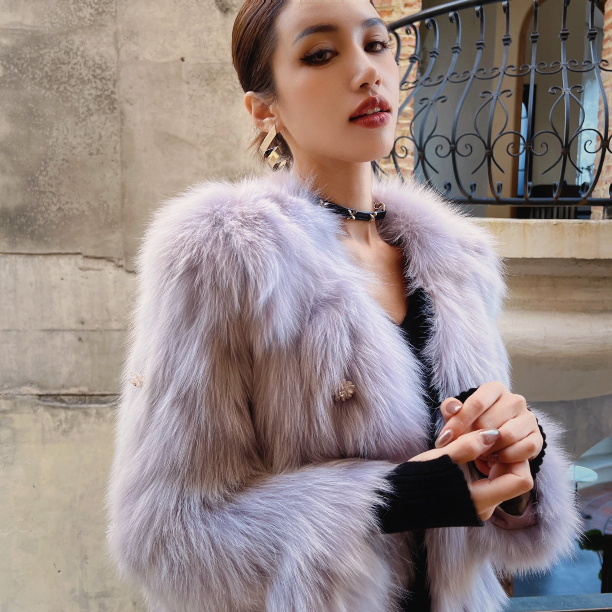A woman wearing a fur coat and holding her hands together, standing in front of a concrete wall.