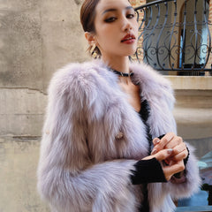 A woman wearing a fur coat and holding her hands together, standing in front of a concrete wall.
