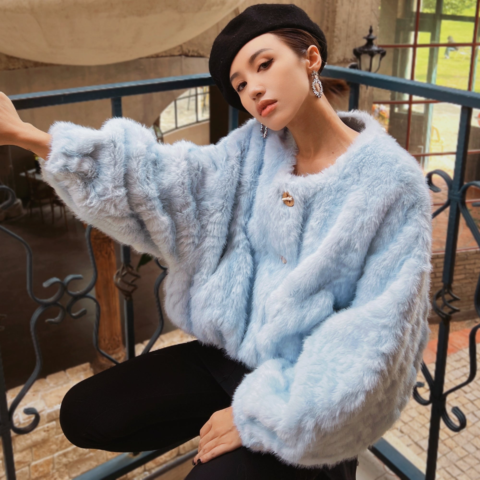 A woman wearing a light blue fur coat and a black beret is sitting on a railing, looking off to the side.