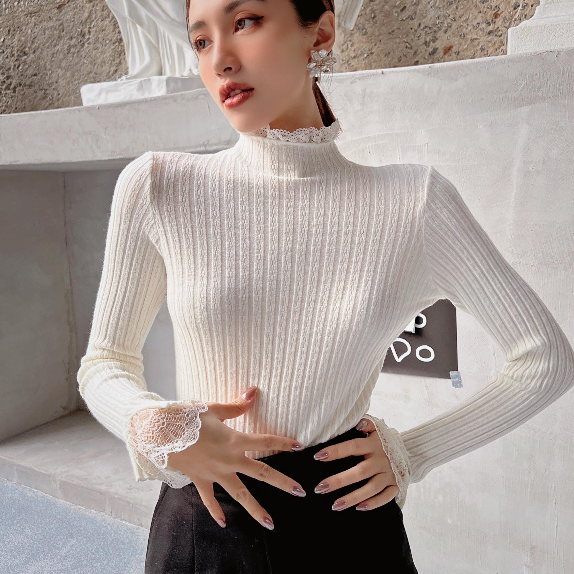 A woman wearing a white turtleneck sweater with lace sleeves, posing in front of a concrete wall.