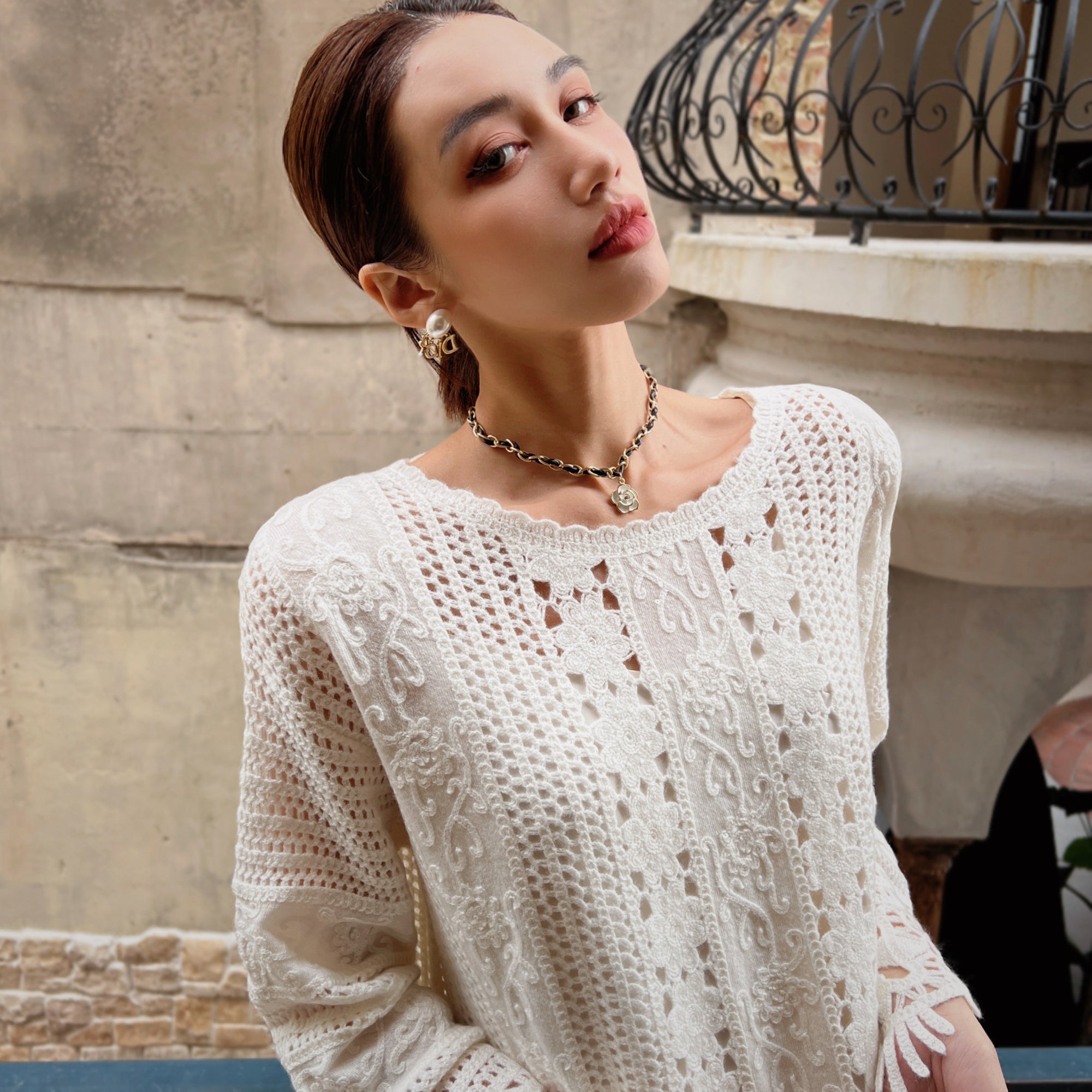 A woman wearing a white lace sweater and earrings stands in front of a stone wall, looking directly at the camera with a serious expression.