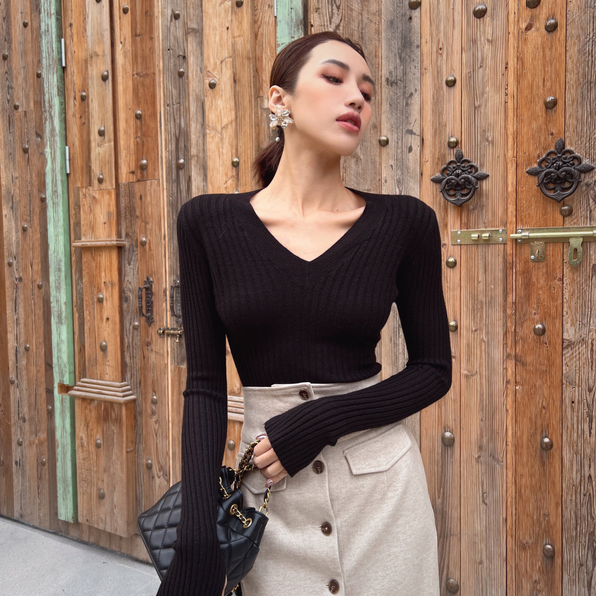 A woman wearing a black sweater and a beige skirt stands in front of a wooden door, holding a black handbag.