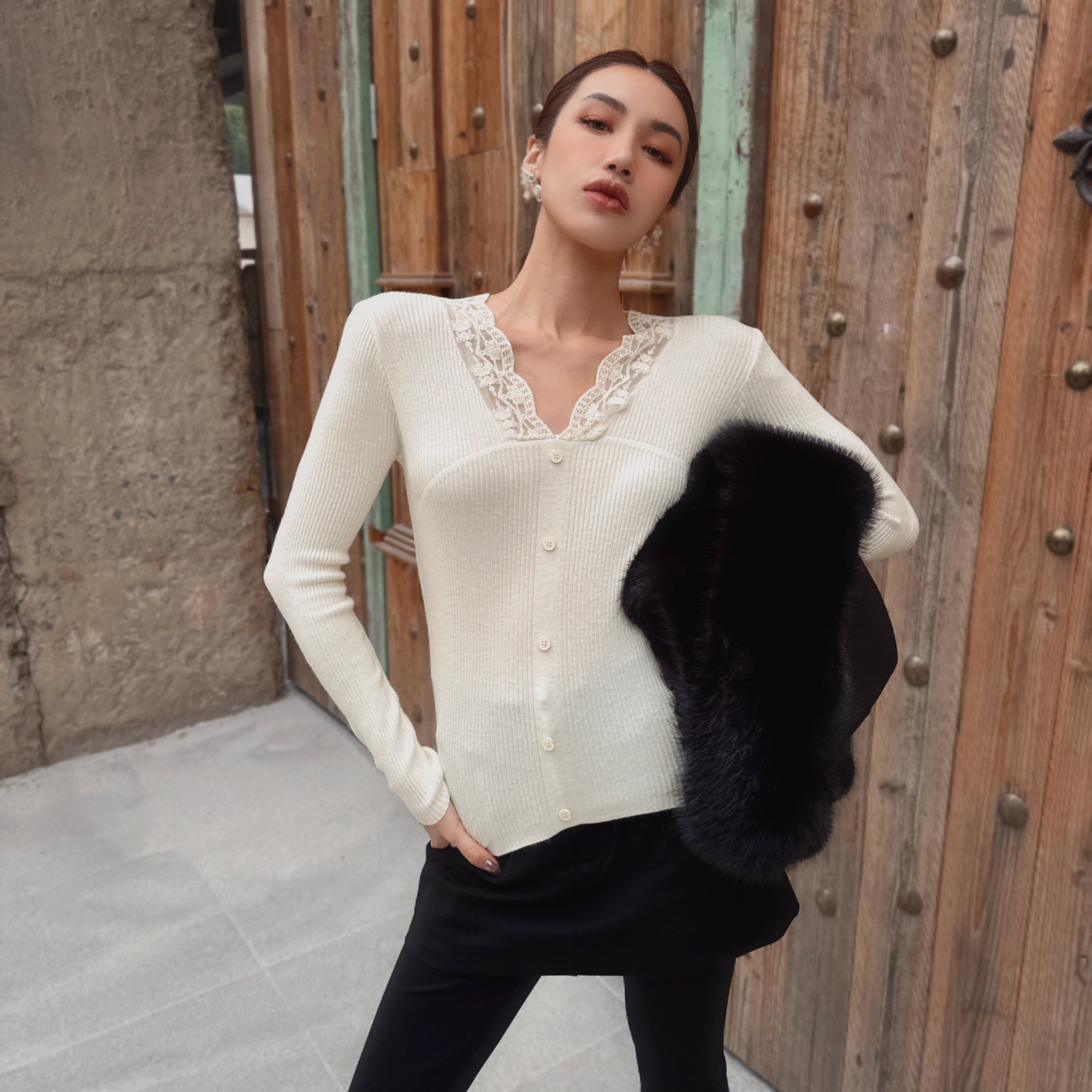 A woman wearing a white cardigan with lace trim and holding a black fur coat stands in front of a wooden wall.