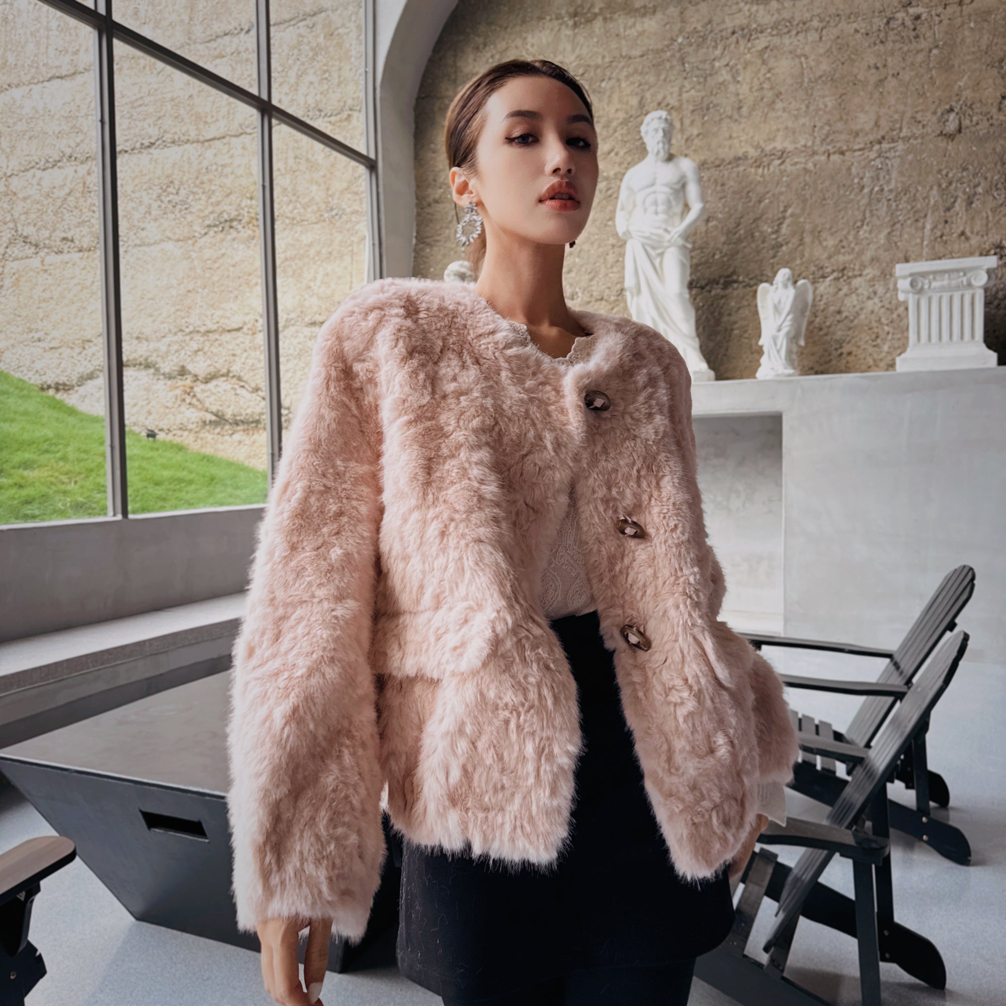 A woman wearing a pink fur coat stands in a room with a window, white statue, and black chairs.