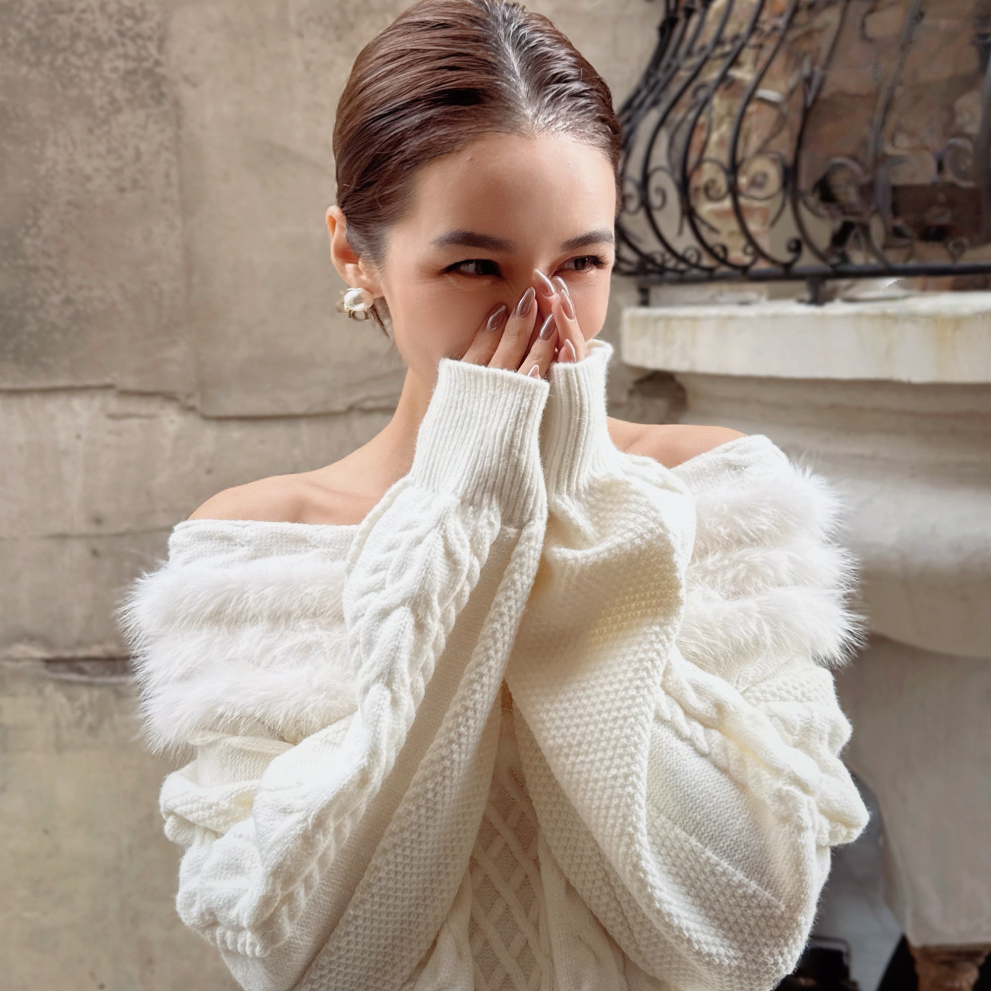 A woman wearing a white sweater with fur trim is covering her face with her hands, with a stone wall and wrought iron gate in the background.