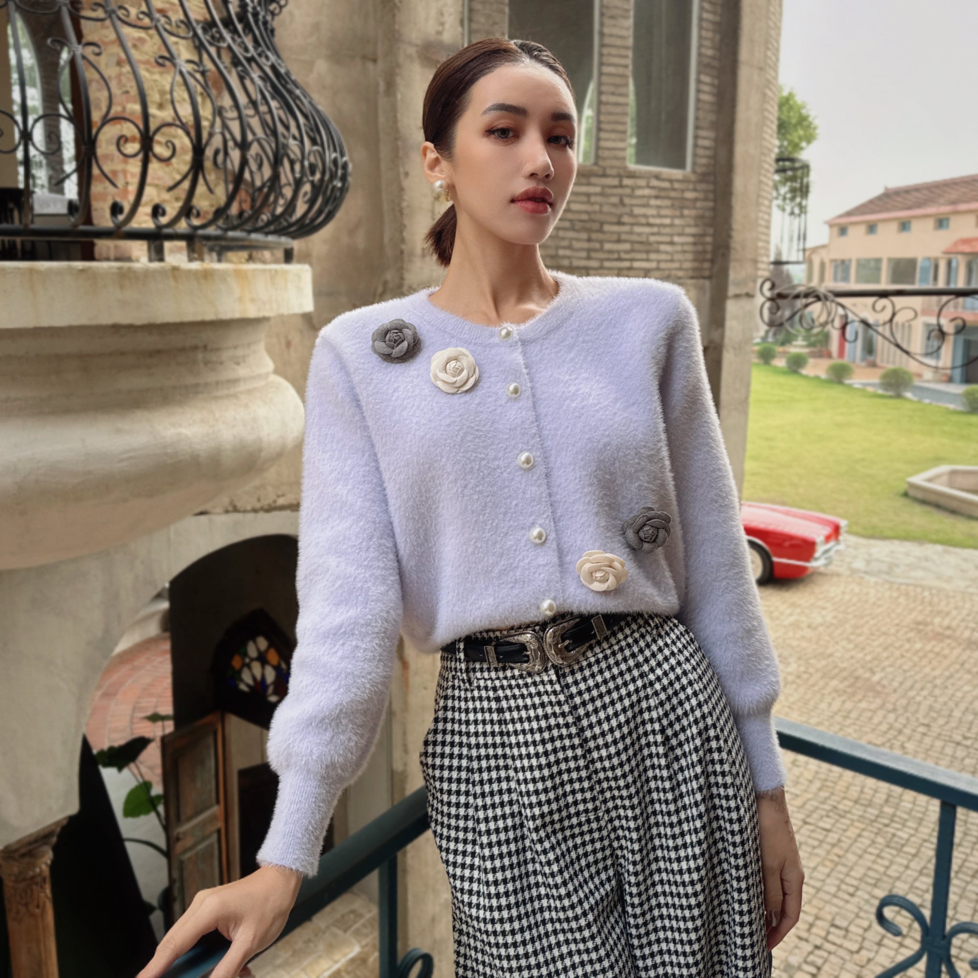 A woman wearing a white cardigan with floral embellishments and a black and white checkered skirt stands in front of a building with a wrought iron balcony.