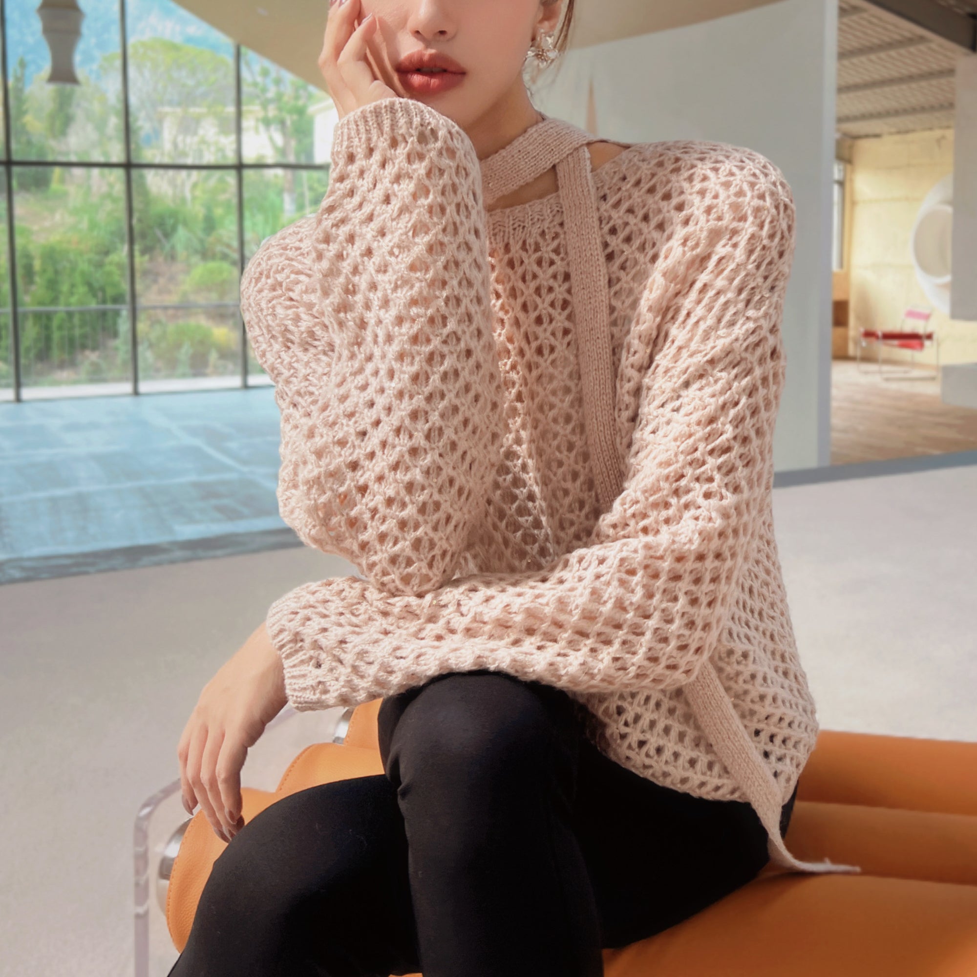 A woman wearing a beige crocheted sweater sits on an orange bench, with a window and white wall in the background.