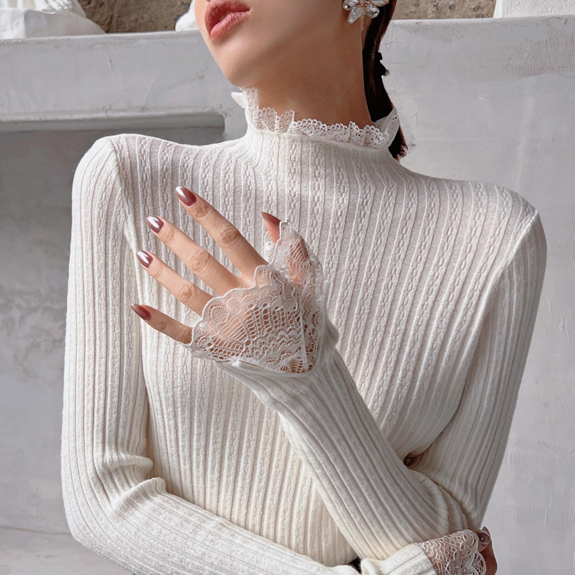 A woman wearing a white lace turtleneck sweater and posing with her hand on her chest.