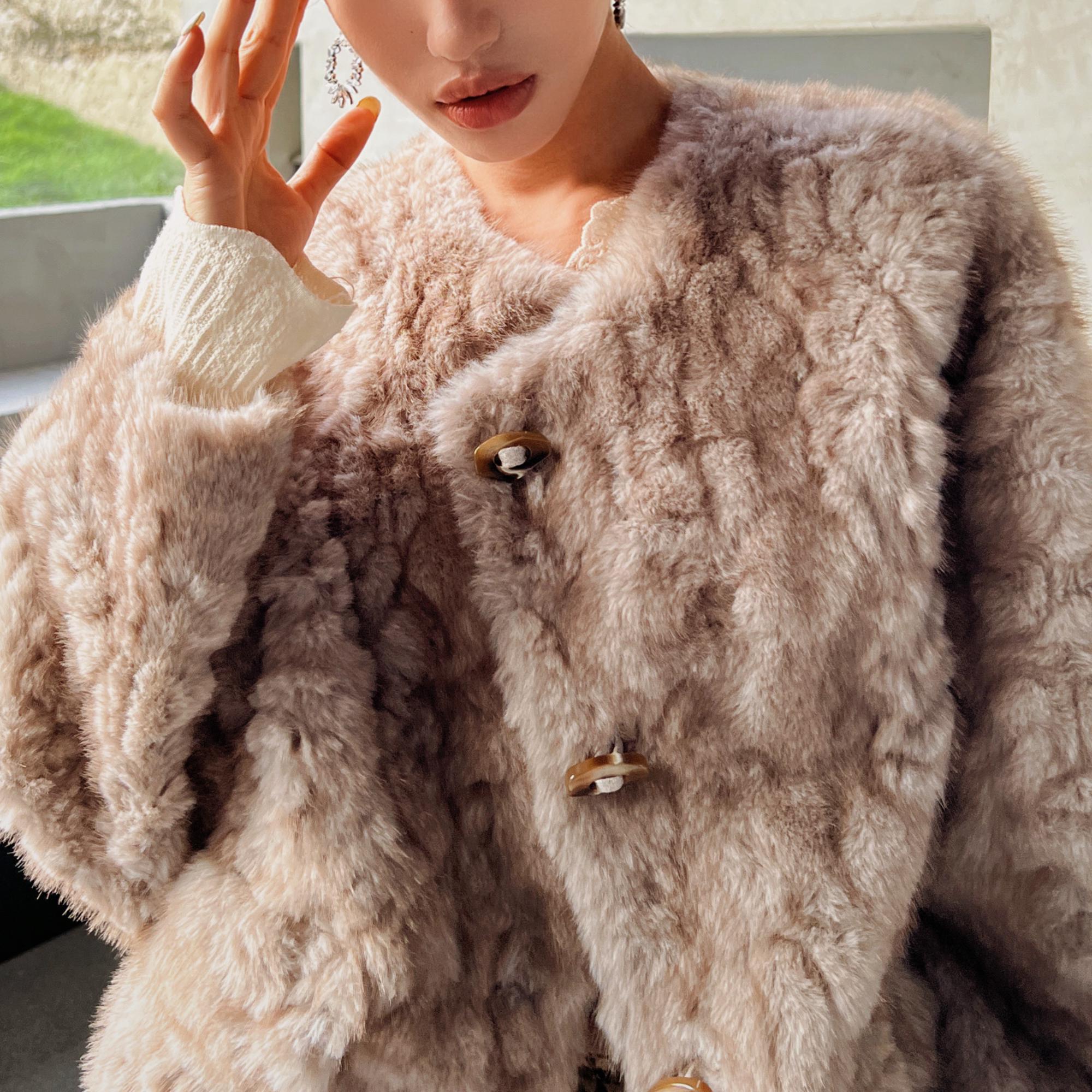 A close-up of a person wearing a beige fur coat, with their hand resting on their face.