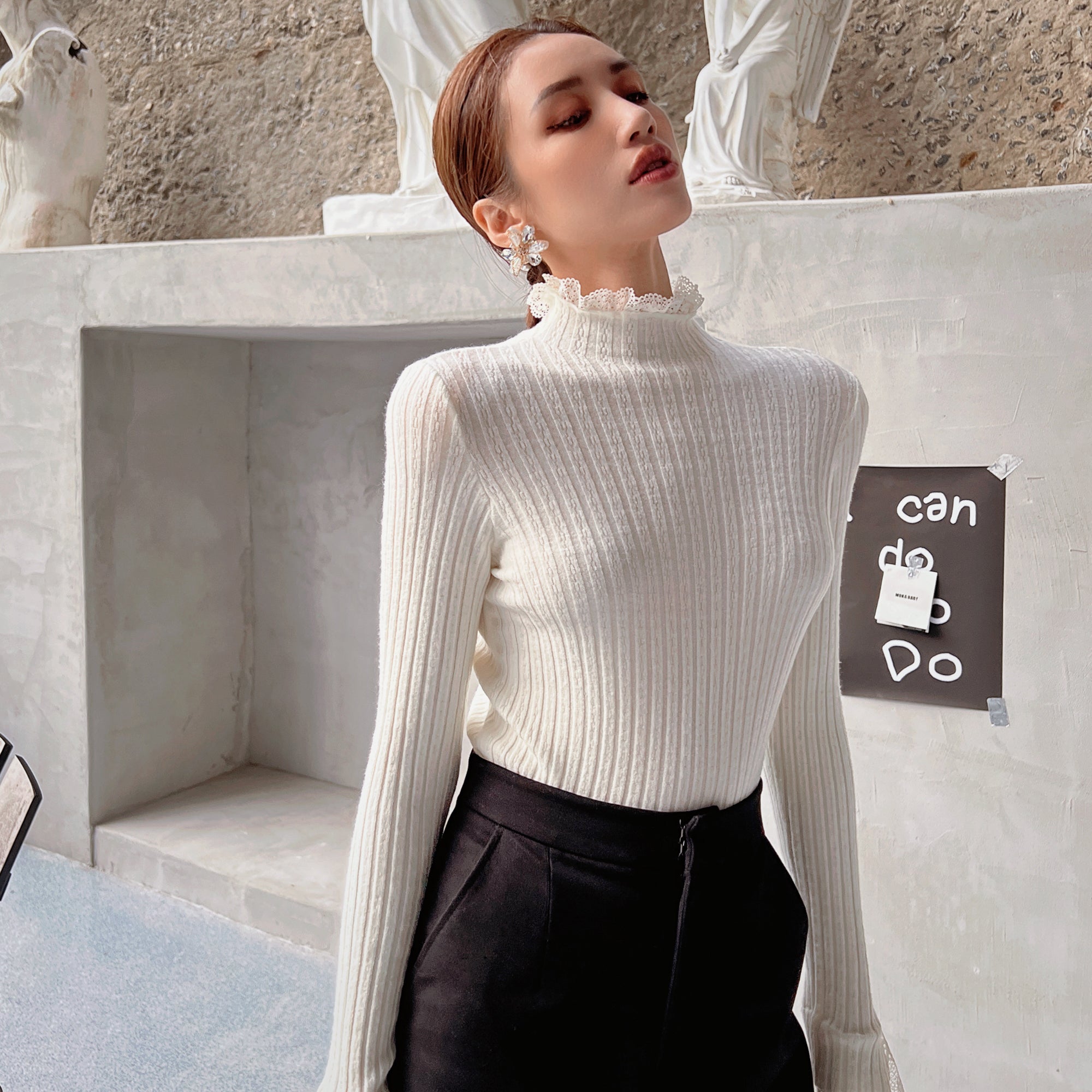 A woman wearing a white sweater and black pants stands in front of a concrete wall with a sign that says "can do".
