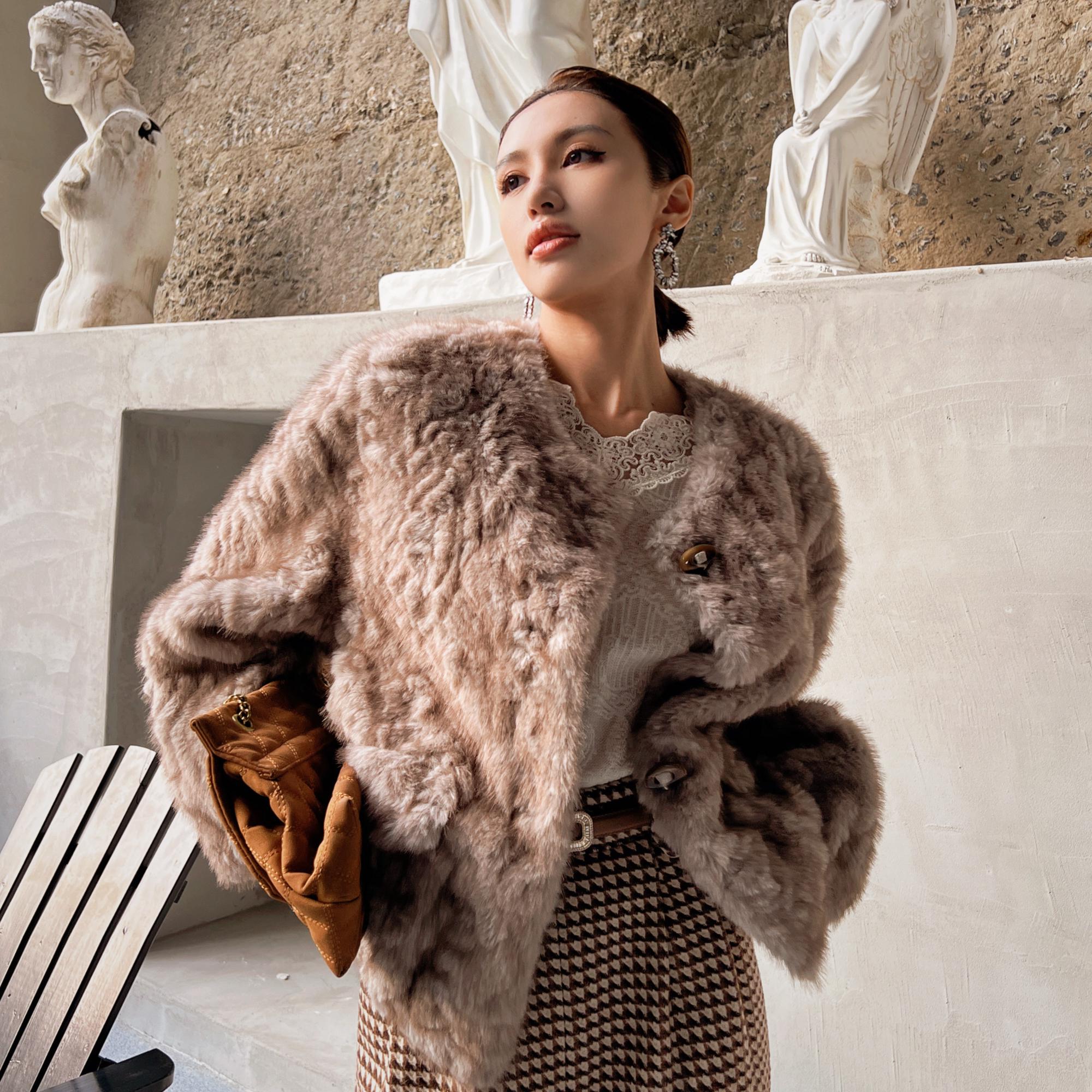 A woman wearing a fur coat and holding a brown purse stands in front of a wall with statues.