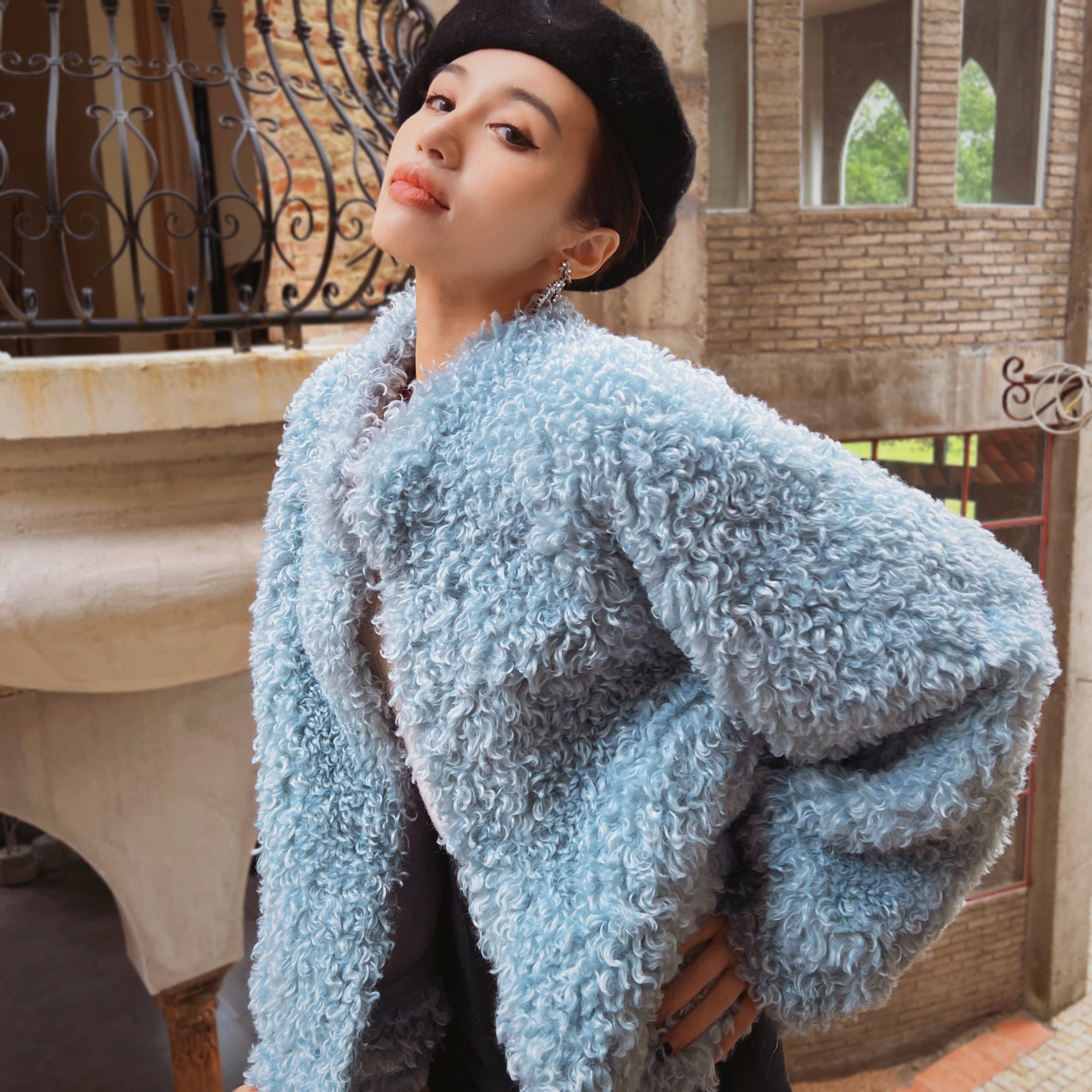 A woman wearing a light blue, fluffy coat stands in front of a brick building with arched windows.